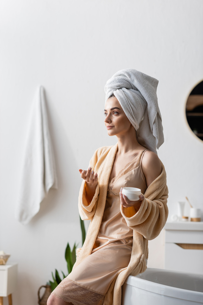junge Frau mit Handtuch auf dem Kopf hält Behälter mit Körperbutter im Badezimmer  - Foto, Bild