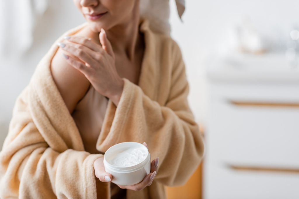 Cropped view of young woman in bathrobe applying cosmetic cream on shoulder in bathroom  - Foto, Imagem