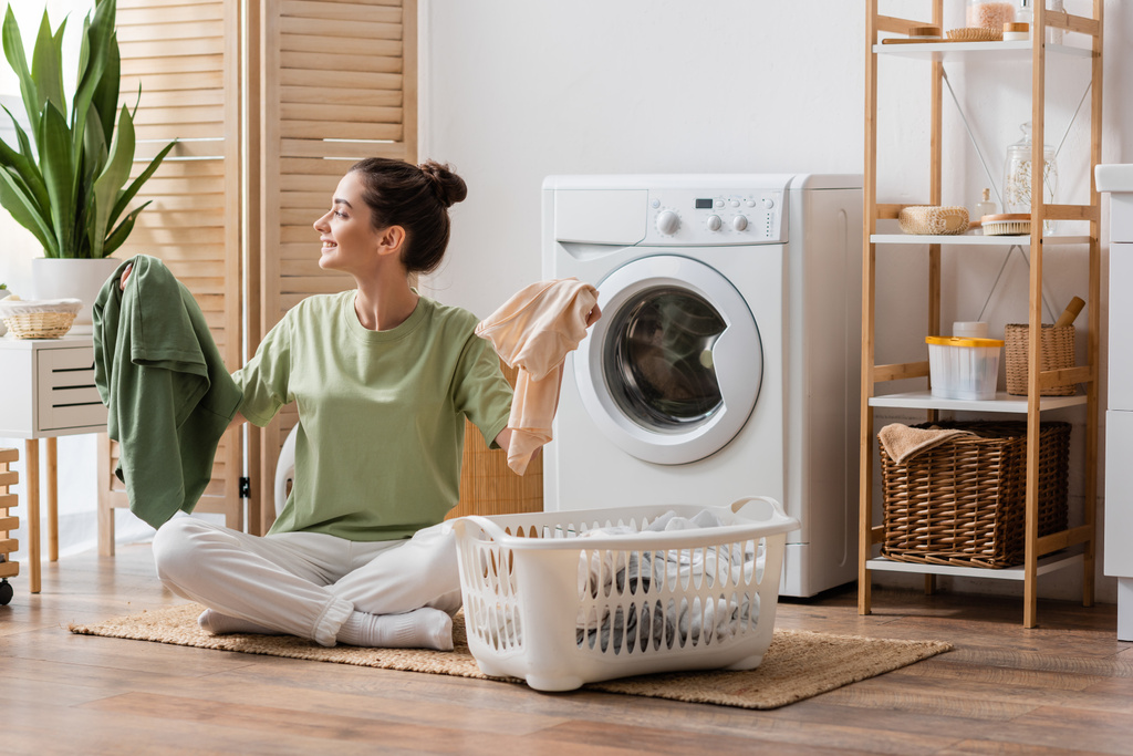 Vista lateral de mujer morena alegre sosteniendo la ropa cerca de la cesta en la sala de lavandería  - Foto, Imagen
