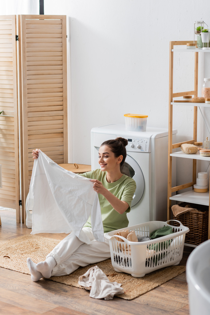 Lächelnde Frau hält Hemd neben Korb und Waschmaschine zu Hause  - Foto, Bild
