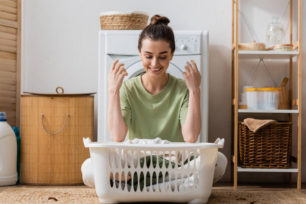 Positive brunette woman looking at basket with clothes in laundry room  - Foto, immagini