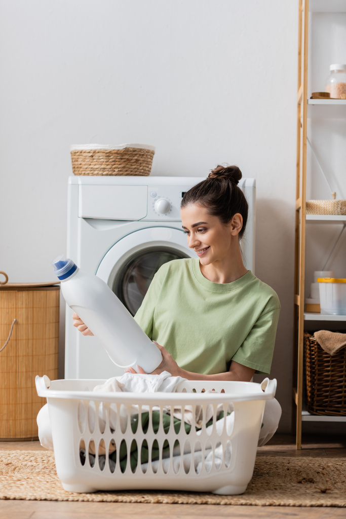 Wesoła brunetka trzyma płyn do prania w pobliżu kosza z ubraniami w pralni  - Zdjęcie, obraz