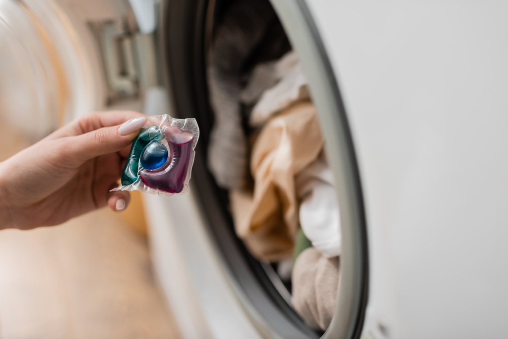 Обрезанный вид женщины, держащей стручок моющего средства возле стиральной машины дома  - Фото, изображение