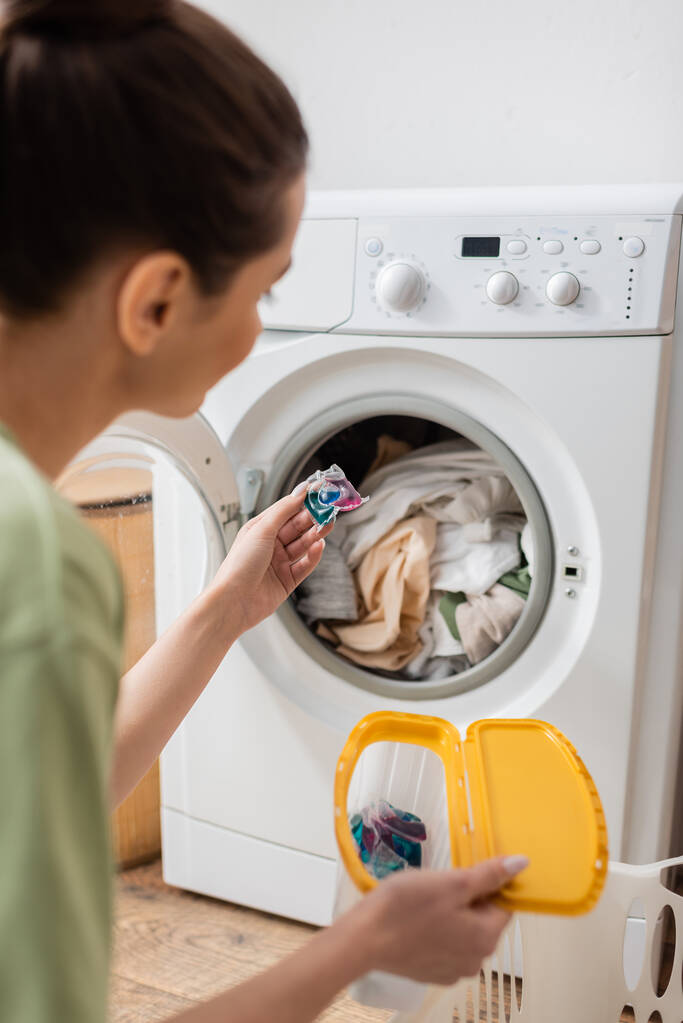 Blurred young woman holding washing capsule near machine in laundry room  - 写真・画像