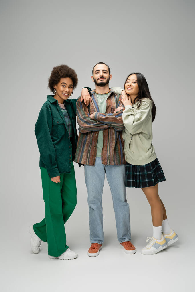 full length of pleased bearded man posing with crossed arms near young multiethnic women on grey background - Foto, afbeelding