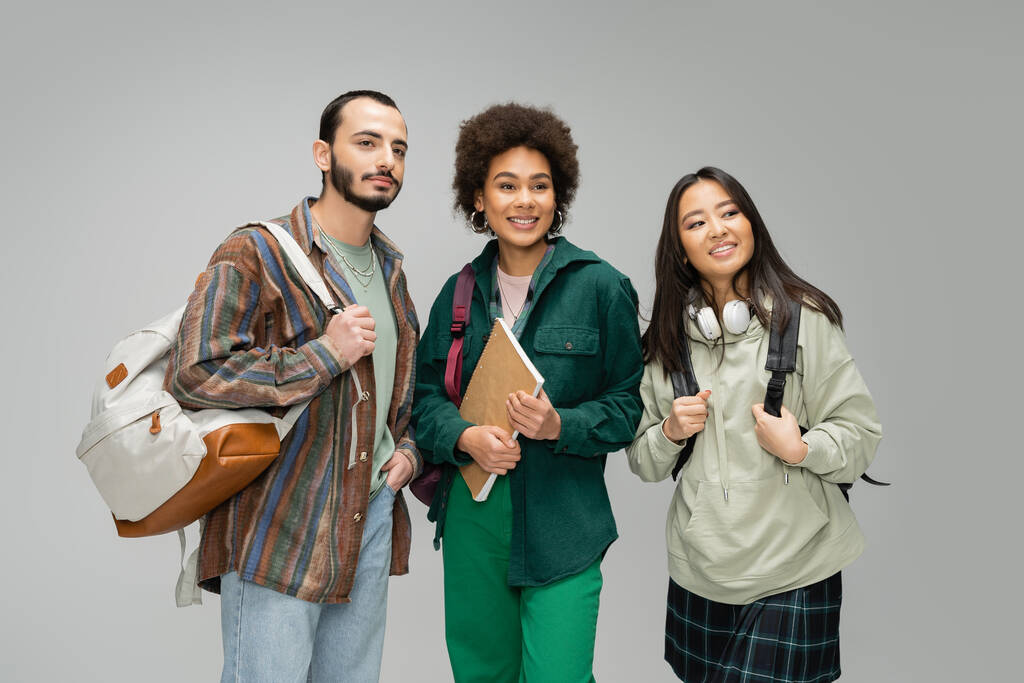 trendy multicultural students with backpacks and notebook smiling and looking away isolated on grey - Fotoğraf, Görsel