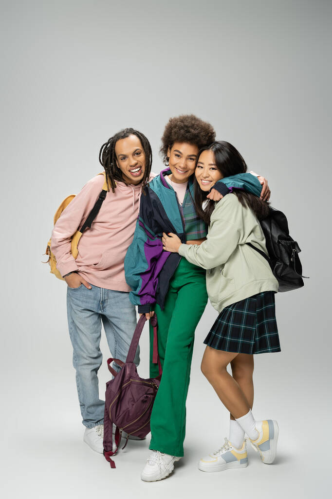comprimento total de estudantes multiétnicos alegres e elegantes com mochilas abraçando em fundo cinza - Foto, Imagem