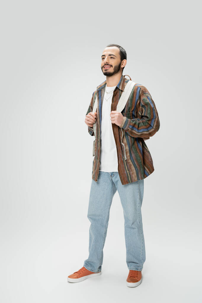 pełna długość uśmiechniętego mężczyzny w niebieskich dżinsach i kolorowa koszula stojąca z plecakiem na szarym tle - Zdjęcie, obraz