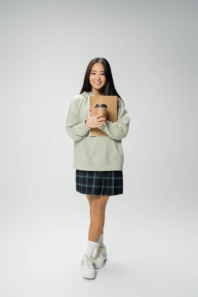 full length of cheerful asian student in checkered skirt and hoodie holding notebook and paper cup on grey background - Fotó, kép