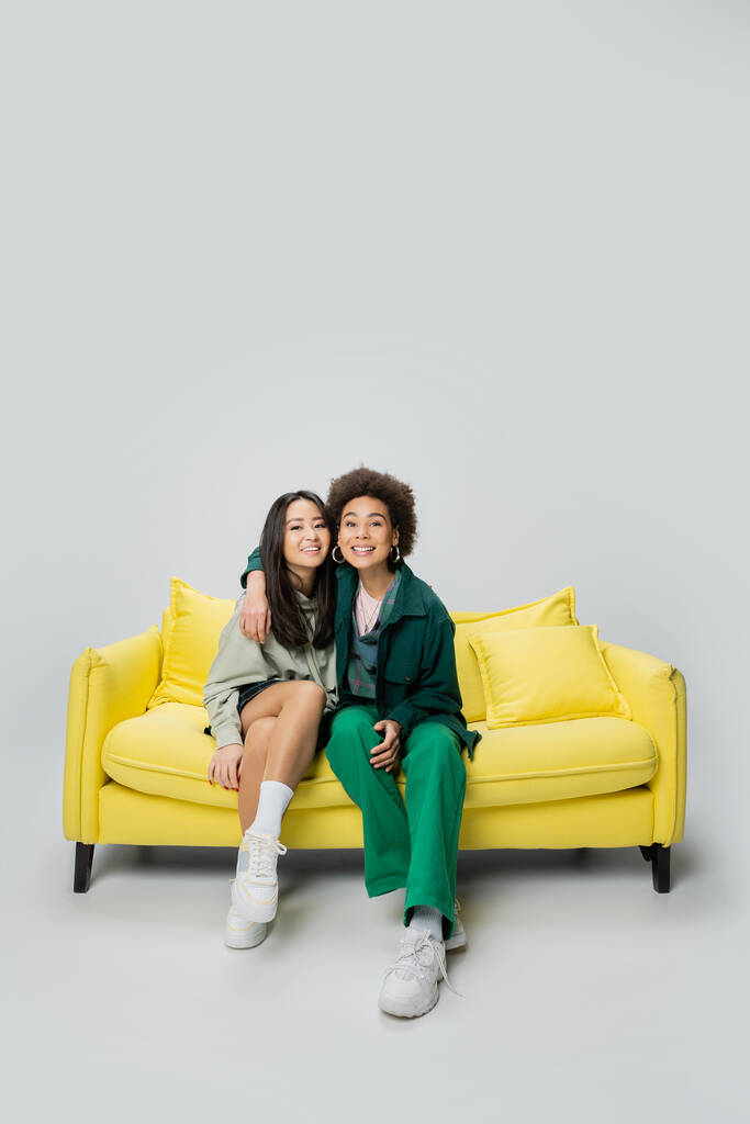 full length of pleased african american woman looking at camera and hugging asian friend on yellow sofa on grey background - Foto, Bild