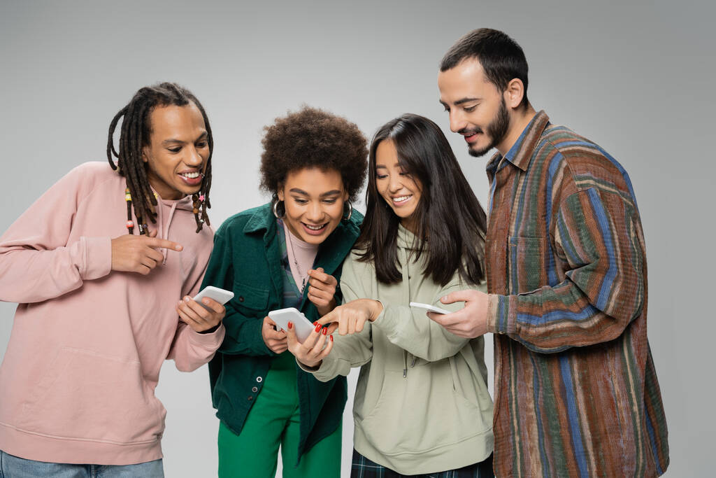 cheerful asian woman pointing at smartphone near trendy multiethnic friends isolated on grey - Foto, Imagem