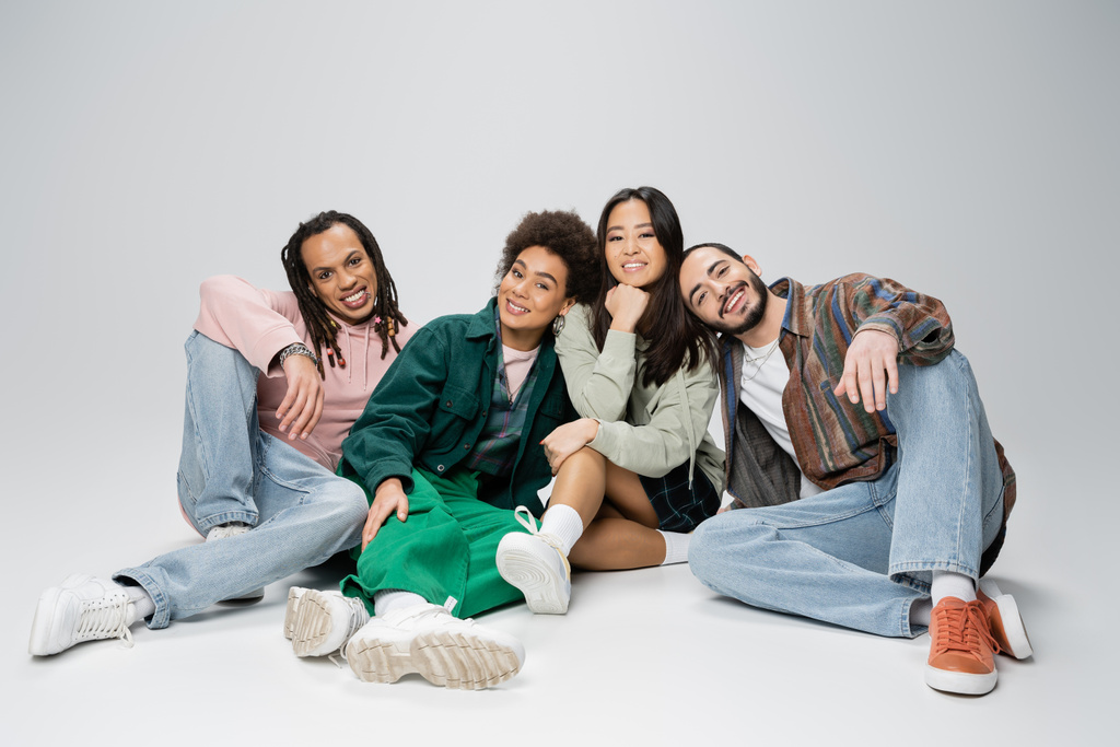 full length of stylish multiethnic friends smiling at camera while sitting on grey background - Fotó, kép