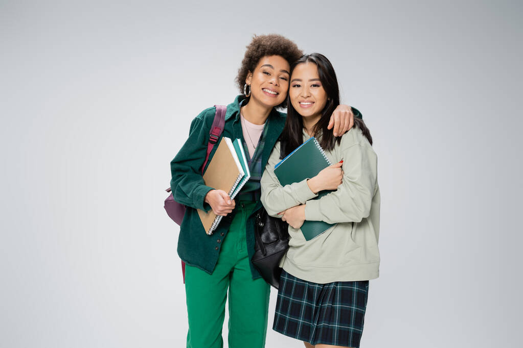 pretty interracial students with copybooks and backpacks smiling at camera isolated on grey - Fotografie, Obrázek