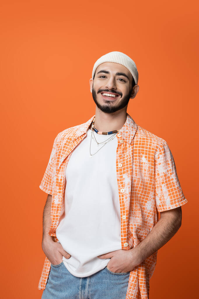 young bearded man in white beanie and beads posing with hands in pockets of jeans isolated on orange - Fotó, kép