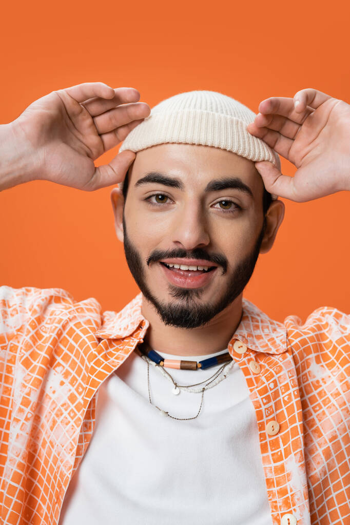 portret młodego brodatego mężczyzny ustawiającego białą czapkę i uśmiechającego się do kamery odizolowanej na pomarańczowo - Zdjęcie, obraz