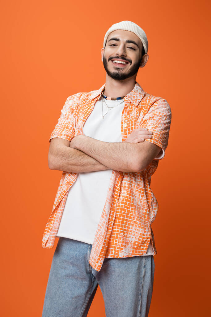hombre barbudo feliz en camisa de moda y gorro blanco posando con brazos cruzados aislados en naranja - Foto, Imagen
