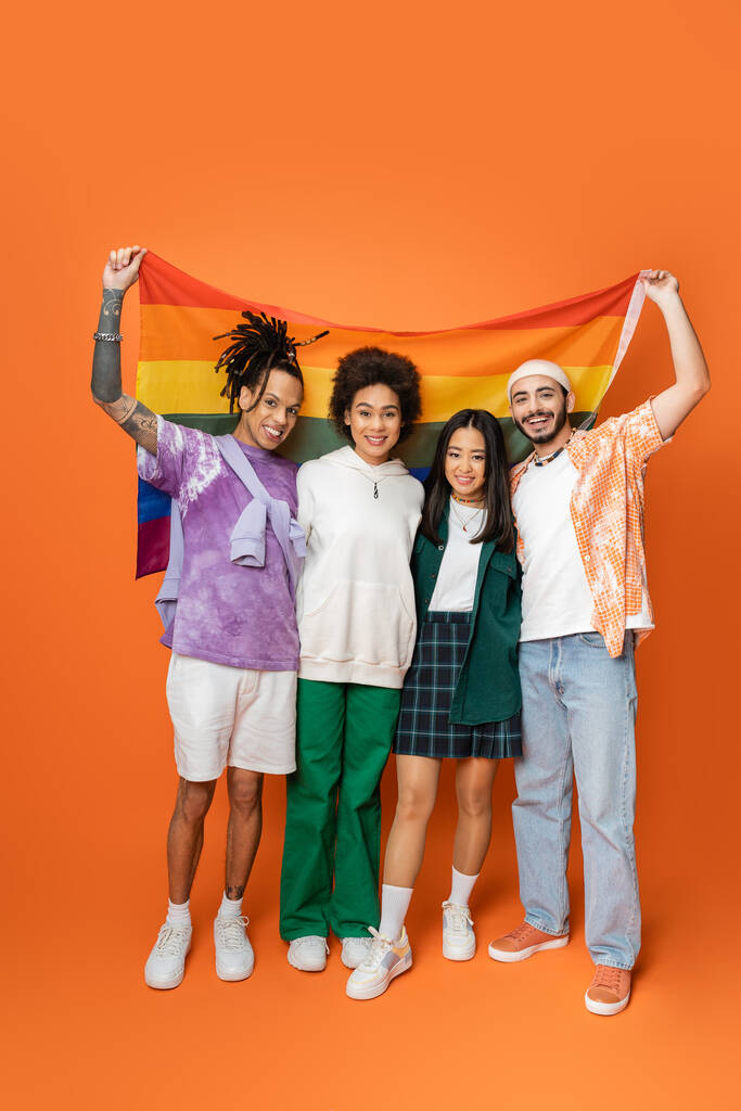 full length of smiling multiethnic friends in trendy clothes holding lgbt flag on orange  - Foto, Imagen