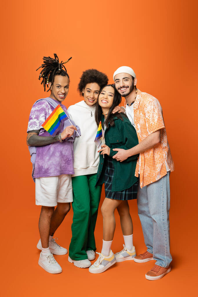 full length of multiracial friends in stylish outfit holding lgbt flags on orange background - Fotó, kép
