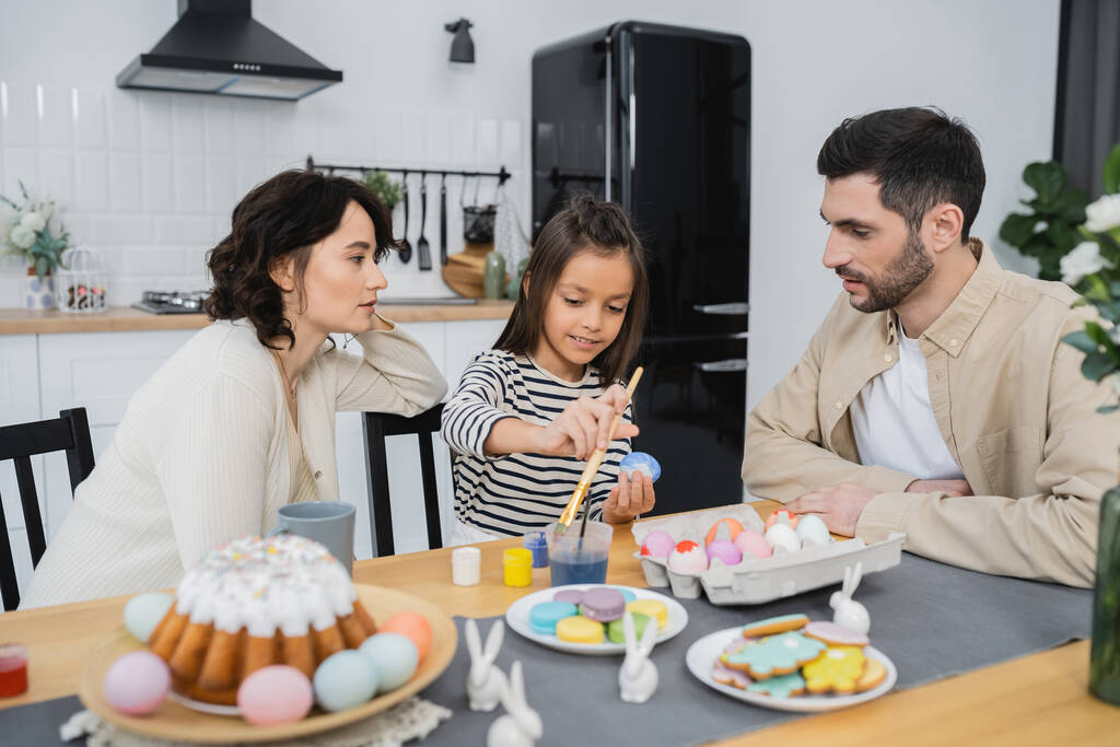 Родители разговаривают рядом с веселой дочерью раскрашивая пасхальные яйца дома  - Фото, изображение