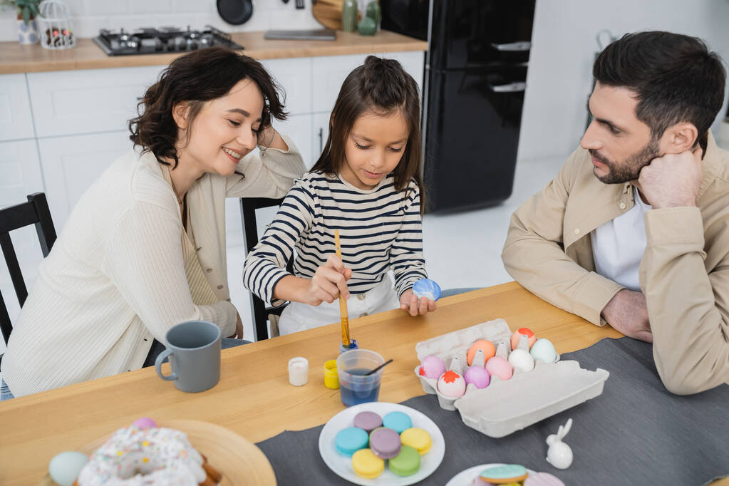 Χαμογελαστή γυναίκα κάθεται κοντά κόρη χρωματισμός πασχαλινά αυγά και ο σύζυγος στο σπίτι  - Φωτογραφία, εικόνα