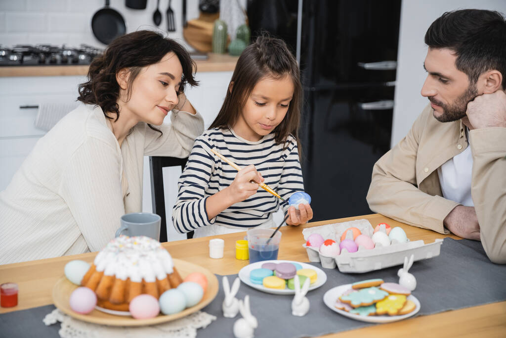 Οικογένεια κάθεται κοντά κόρη χρωματισμός αυγά Πάσχα κοντά στο φαγητό στο σπίτι  - Φωτογραφία, εικόνα