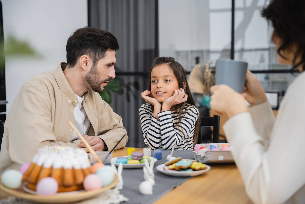 Hombre hablando con la hija sonriente cerca de la comida de Pascua en la cocina en casa  - Foto, Imagen