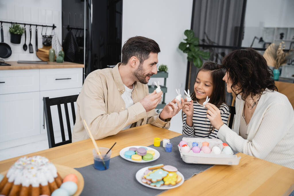 Positiiviset vanhemmat ja lapsi, joilla on pääsiäiskaneja lähellä kakkua ja munia kotona  - Valokuva, kuva