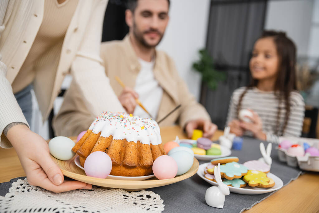 Woman putting plate with Easter cake on table near blurred family at home  - Fotó, kép