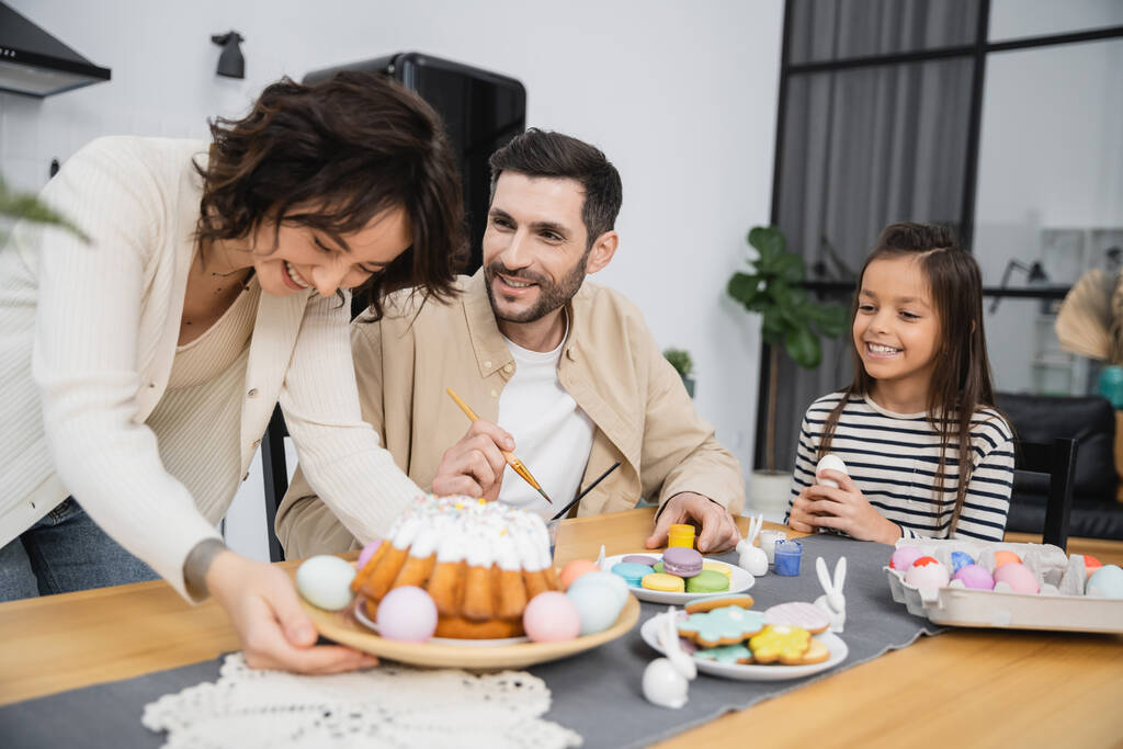 Smiling man and girl coloring Easter eggs near woman with cake at home  - Photo, Image