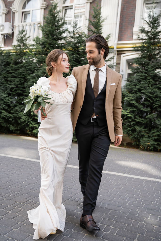 full length of gorgeous bride in white dress holding wedding bouquet while standing with bearded groom on street  - Foto, Bild