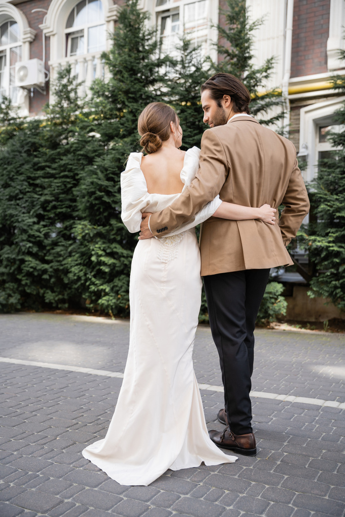 achteraanzicht van bruid in witte jurk knuffelen met bebaarde bruidegom op straat  - Foto, afbeelding