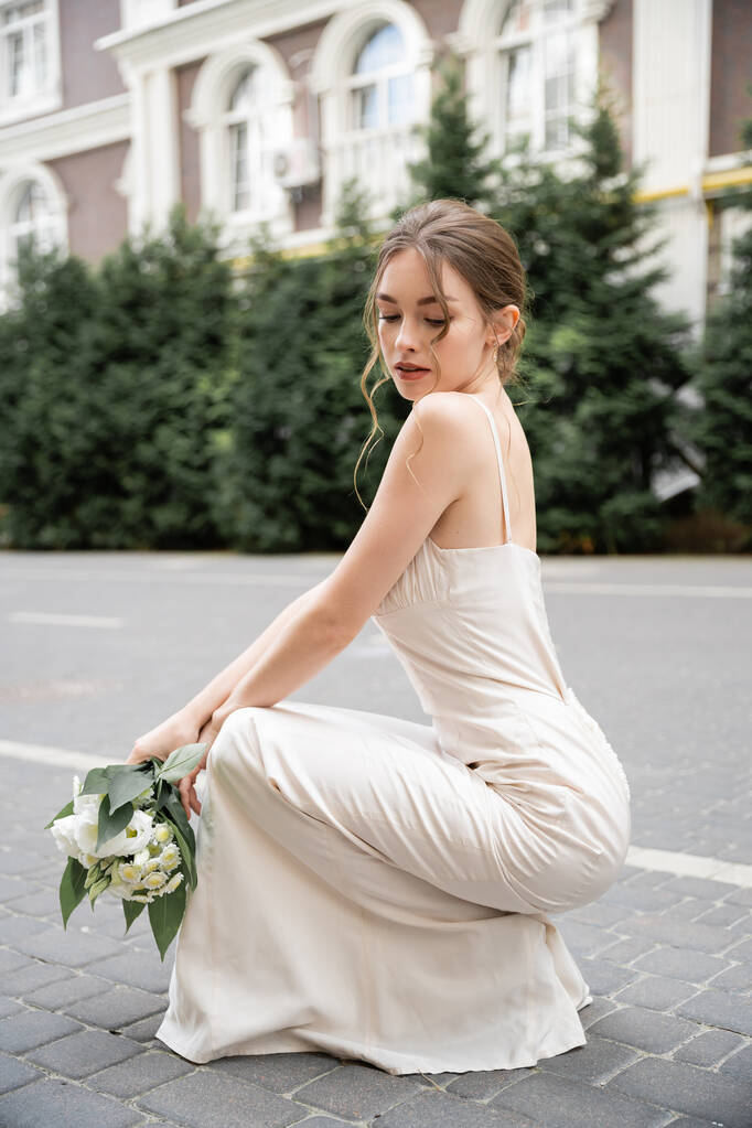 young bride in wedding dress holding bouquet with flowers while sitting outside  - Foto, Imagen