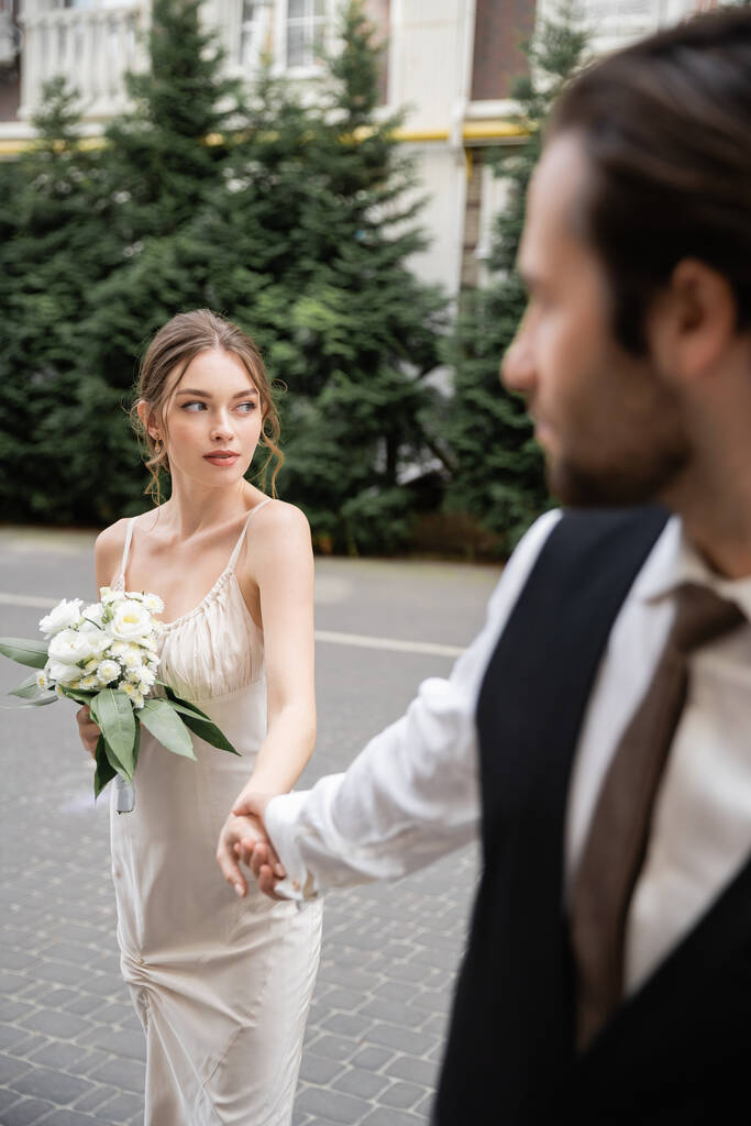 blurred groom in vest holding hand of gorgeous bride in white dress with wedding bouquet  - Fotoğraf, Görsel
