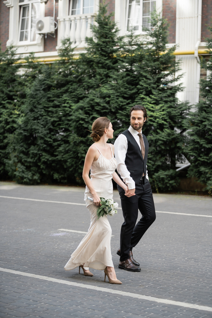 повна довжина чоловіка в костюмі тримає руку нареченої в білій сукні з букетом квітів
  - Фото, зображення
