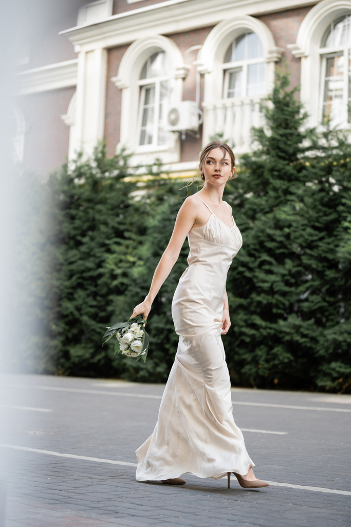 volle Länge der jungen Braut in weißem Kleid mit Hochzeitsstrauß aus blühenden Blumen und draußen gehen  - Foto, Bild