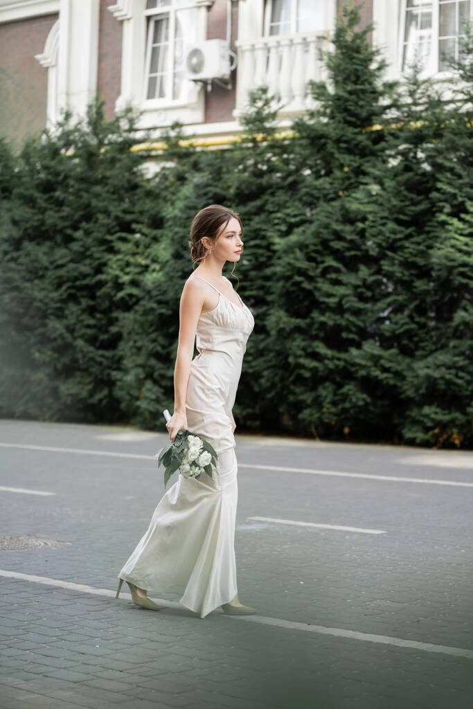 full length of young woman in white dress holding wedding bouquet of flowers and standing outside  - Foto, immagini