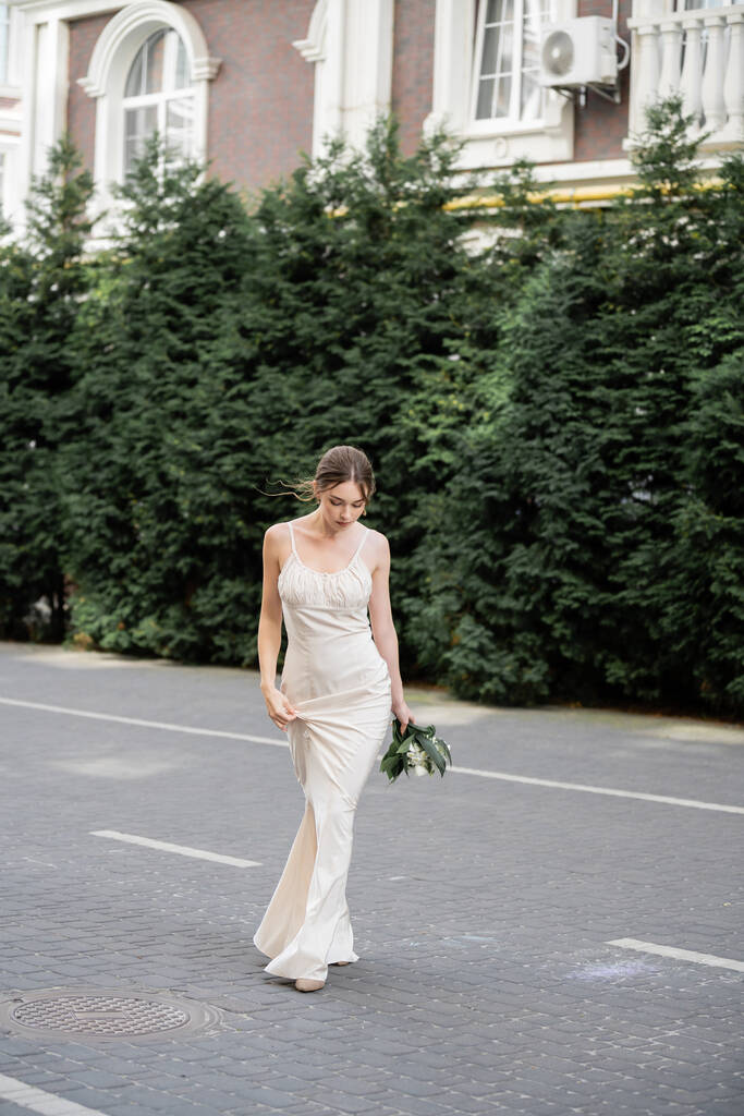 full length of young bride in white dress holding wedding bouquet of flowers and walking on street  - Foto, Bild