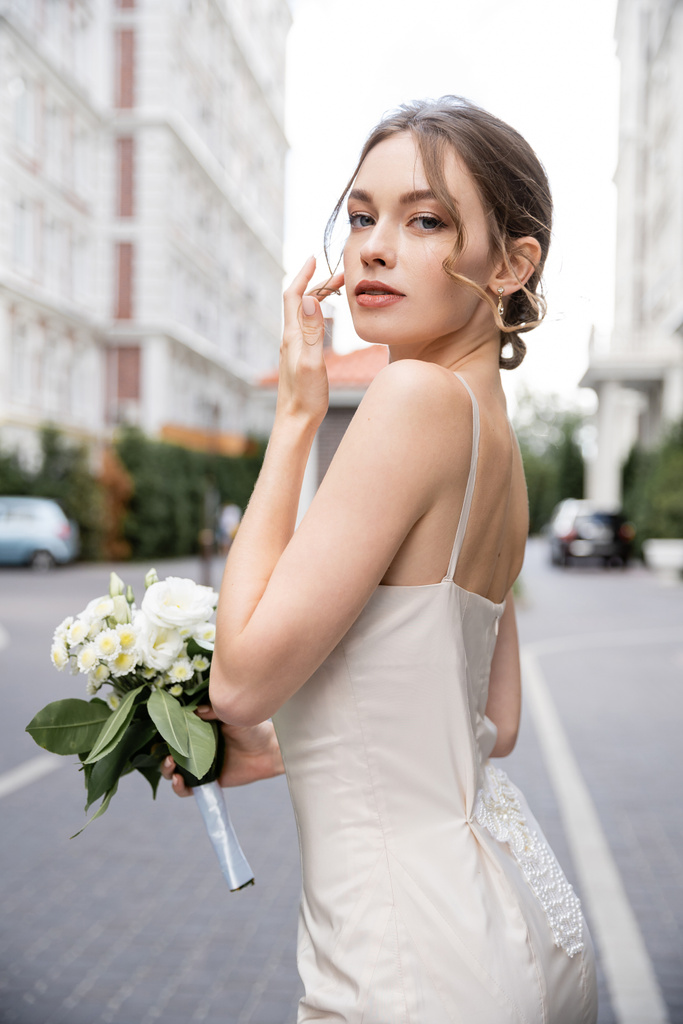 νεαρή γυναίκα σε λευκό φόρεμα κρατώντας γαμήλιο μπουκέτο και κοιτάζοντας την κάμερα  - Φωτογραφία, εικόνα