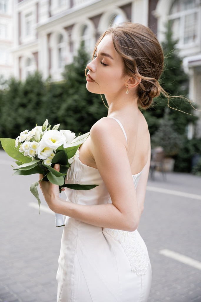 молода наречена в білій сукні стоїть з закритими очима і тримає весільний букет
  - Фото, зображення
