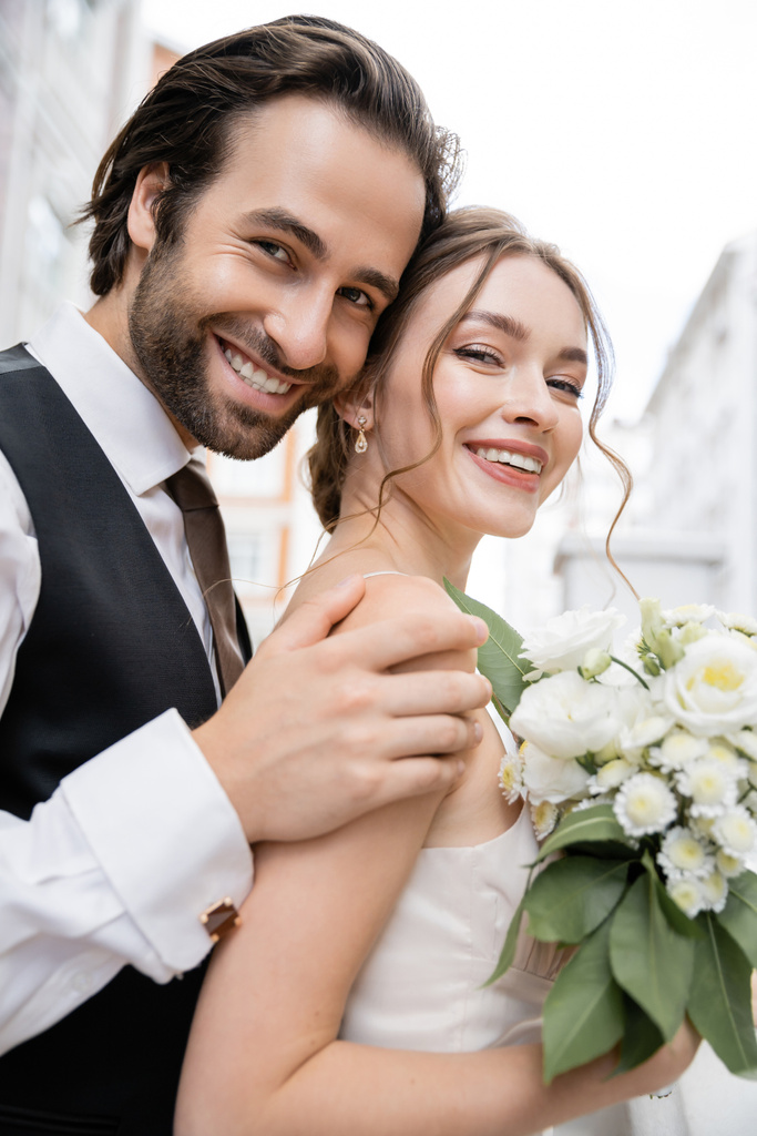 portrait of happy groom with beard hugging cheerful bride holding wedding bouquet  - Foto, afbeelding