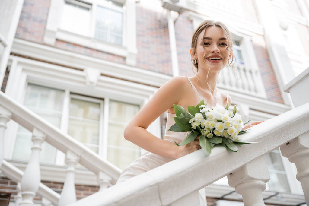 vista de ángulo bajo de la novia feliz en vestido de novia con ramo de celebración y mirando a la cámara cerca de casa  - Foto, Imagen