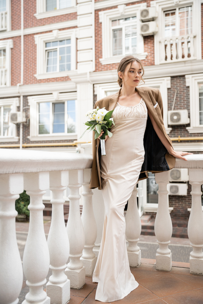 full length of young bride in wedding dress and beige blazer holding bouquet near house  - Photo, image