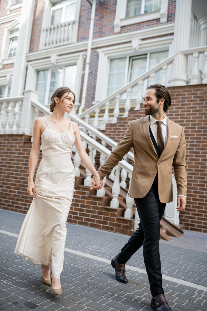 full length of happy newlyweds smiling and holding hands while walking on street  - Fotoğraf, Görsel