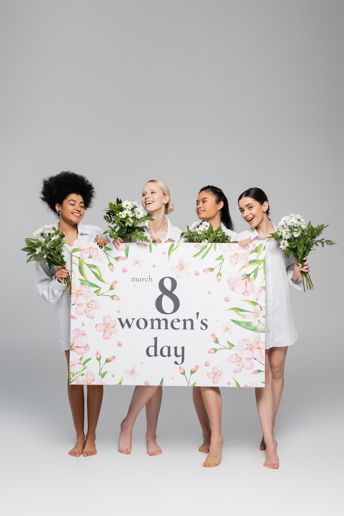 volledige lengte van barefoot multi-etnische vrouwen met bloemen en bordje met vrouwen dag belettering op grijze achtergrond - Foto, afbeelding
