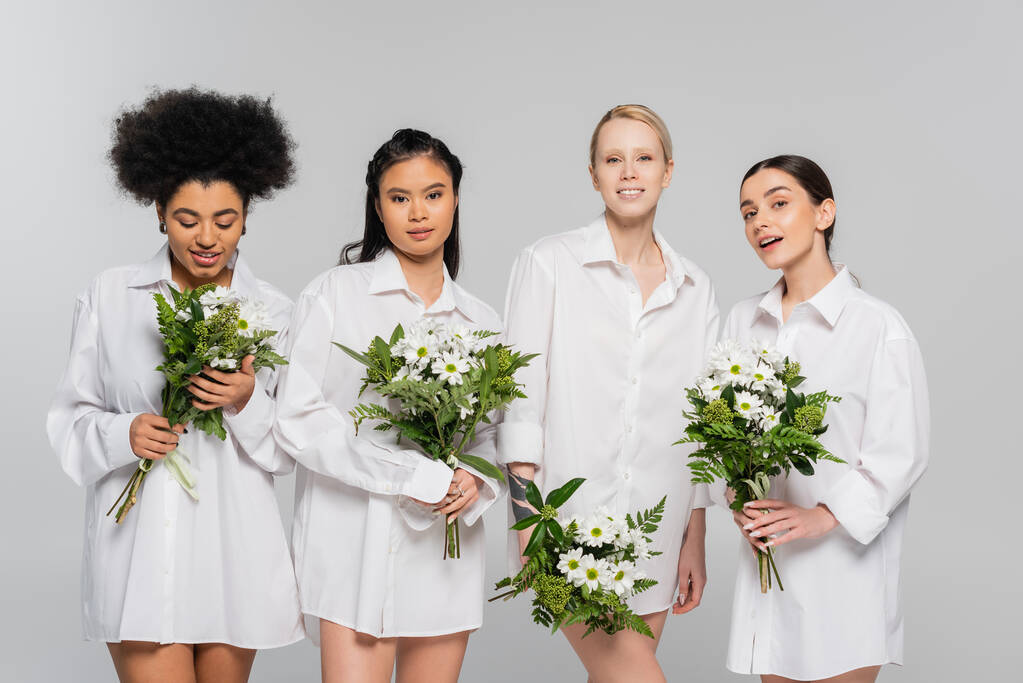 positive multicultural women in white shirts holding bouquets isolated on grey - Foto, imagen