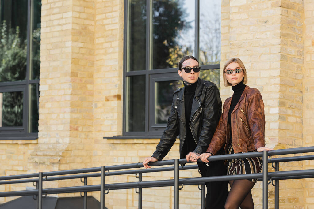young couple in trendy sunglasses and leather jackets standing together outdoors  - Zdjęcie, obraz