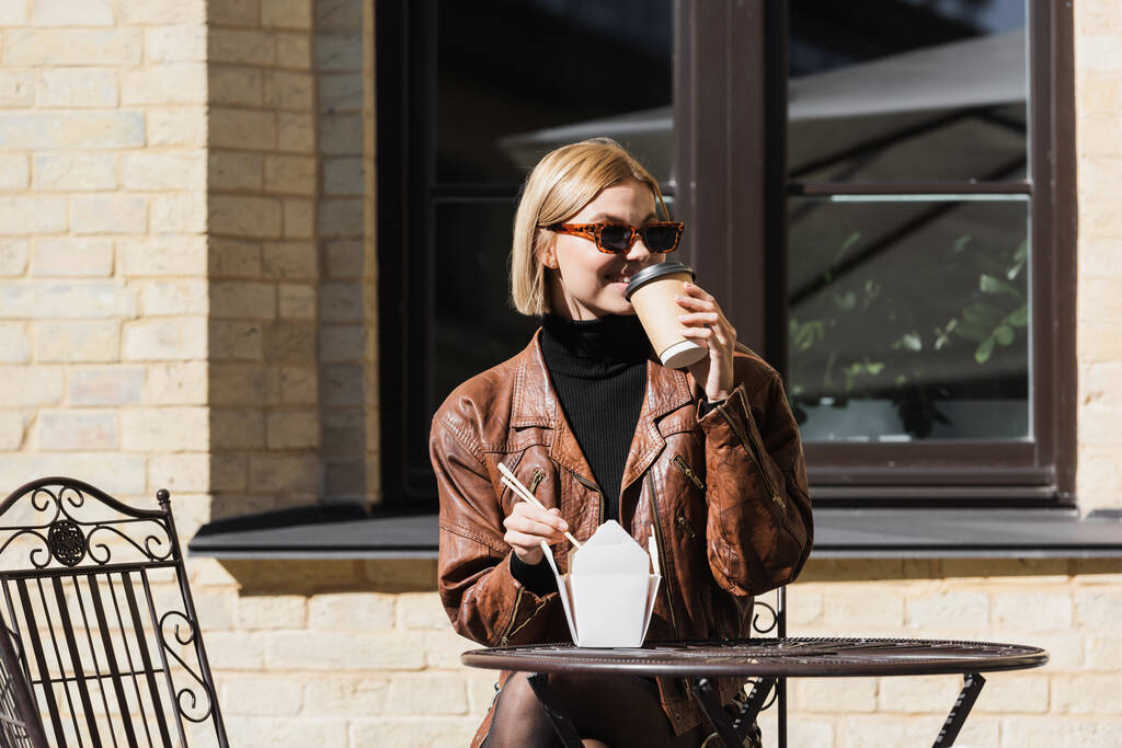 glückliche blonde Frau mit Sonnenbrille hält Essstäbchen in der Nähe von Imbissbuden mit asiatischem Essen und trinkt Kaffee  - Foto, Bild