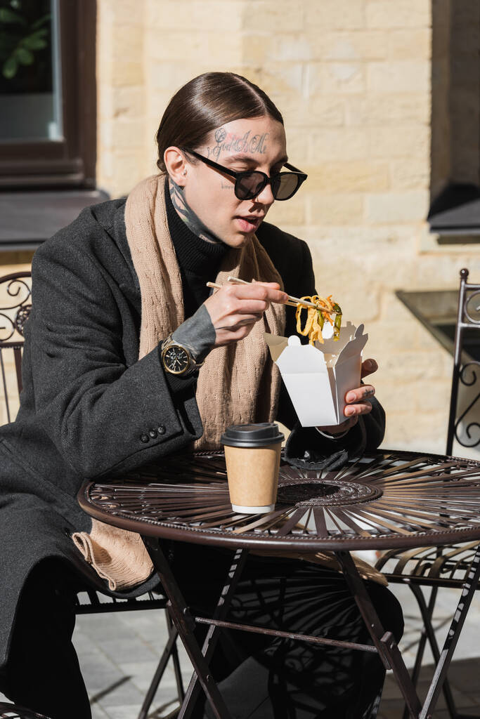 tattooed man in stylish sunglasses holding chopsticks while eating asian food near paper cup on bistro table  - Foto, Imagem