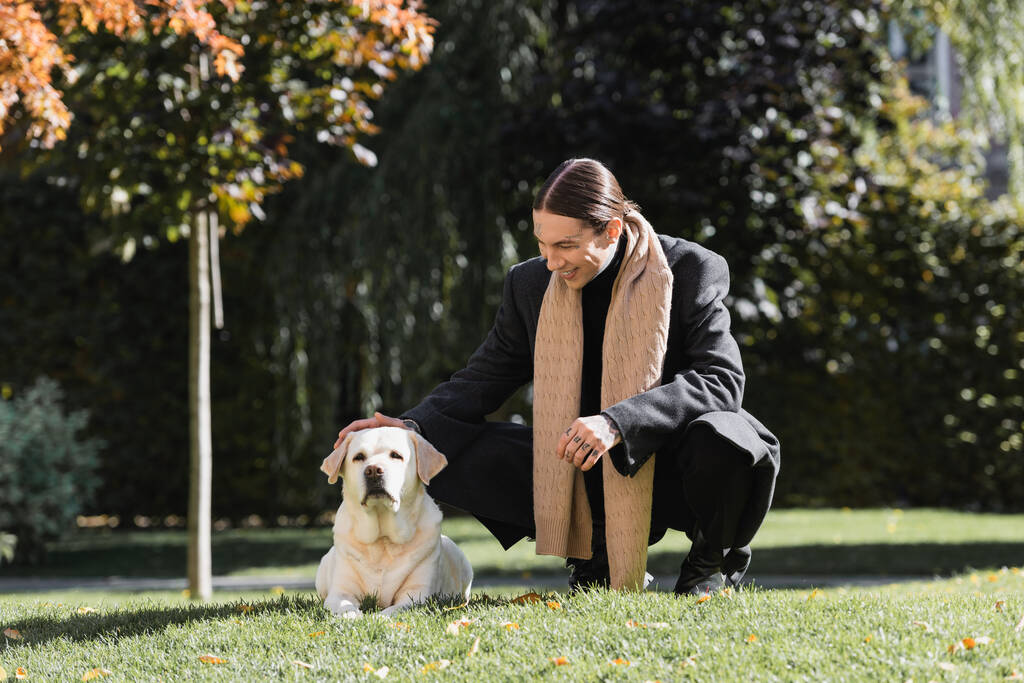 full length of tattooed man in stylish coat and scarf smiling while cuddling labrador in park  - Foto, Imagen