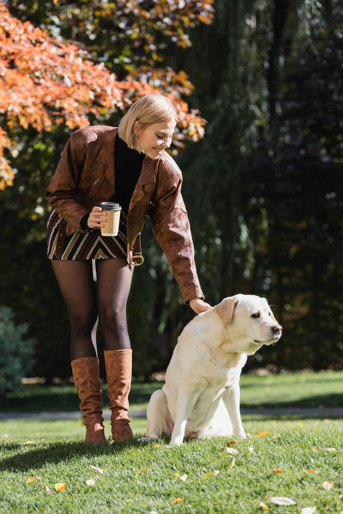 full length of happy blonde woman holding paper cup and cuddling labrador in park  - Fotografie, Obrázek